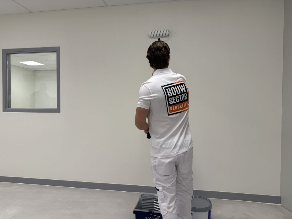 Schilder saust een witte wand met de roller naast een binnenraam, verfbak aan zijn voeten