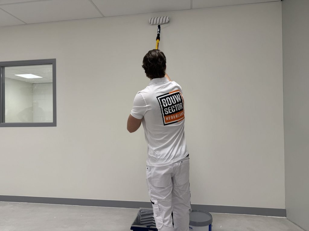 Schilder in wit werktenue rolt de bovenkant van een lichte muur, met verfbak en emmer op de vloer