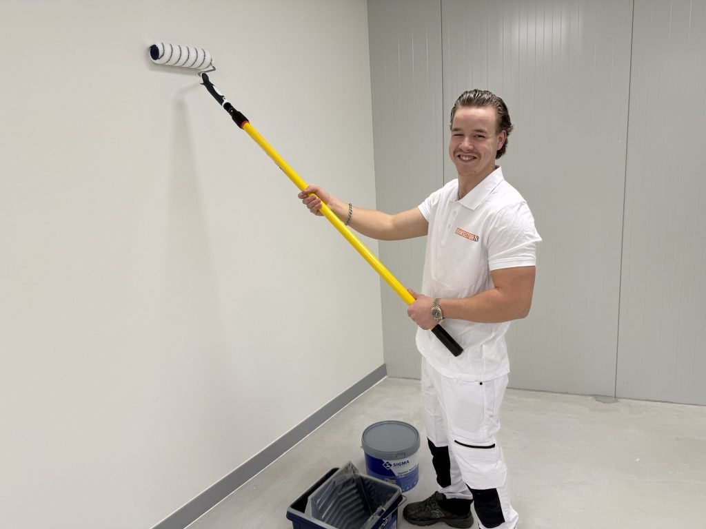 Lachende schilder in witte werkkleding rolt een witte muur, met een emmer Sigma-verf op de vloer