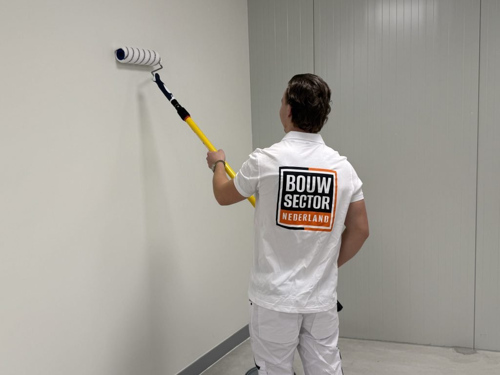 Schilder met roller aan een gele verlengsteel saust een lichte muur in een lege ruimte