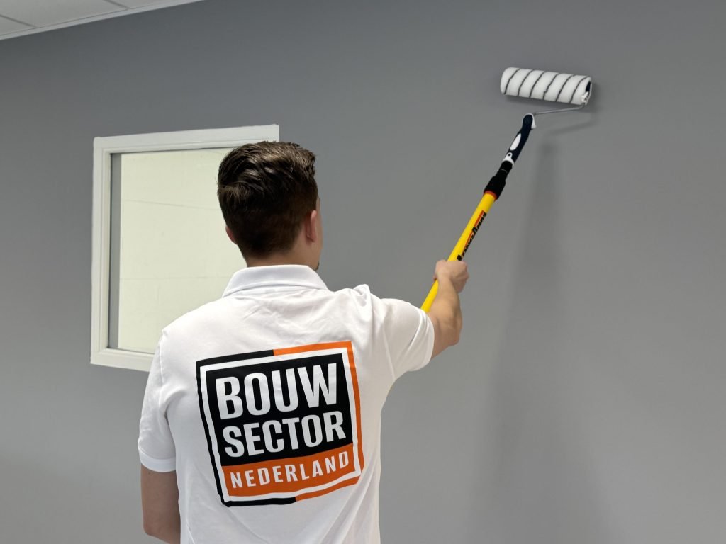 Schilder van Bouwsector Nederland rolt grijze muurverf op een wand naast een raam met wit kozijn