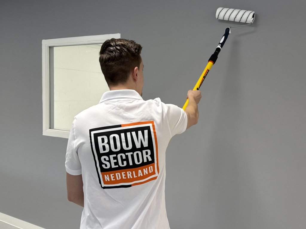 Schilder met Bouwsector Nederland-logo op de rug rolt grijze muurverf naast een raamkozijn