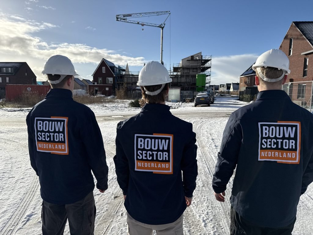 Drie vakmensen van Bouwsector Nederland in donkerblauwe werkjas met bouwhelm, van achteren gezien bij nieuwbouwhuizen en een bouwkraan